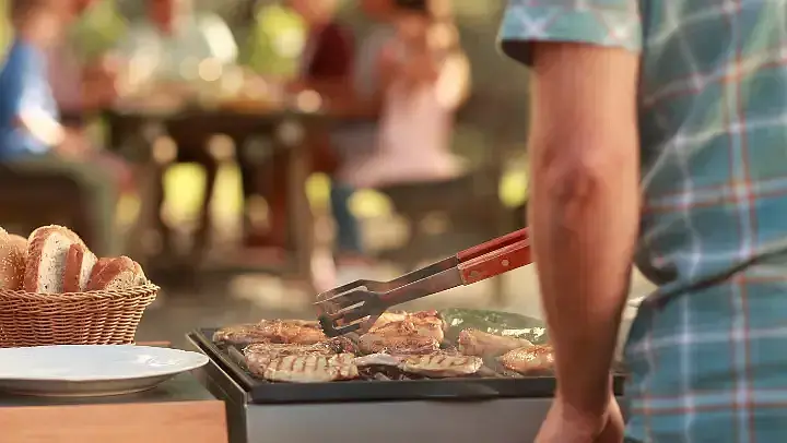 Eine Person grillt Fleisch auf einer Grillplatte. Im unscharfen Hintergrund sitzen mehrere Menschen gemeinsam an einem Tisch und essen. Die Szene vermittelt eine gesellige Grillfeier im Freien.