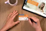 Tisch in der Draufsicht, darauf zentral zwei Hände, die einen Organspendeausweis ausfüllen. Außerdem im Anschnitt ein Tablett mit der Webseite organspende-info.de und ein halb gefüllte Kaffeetasse 