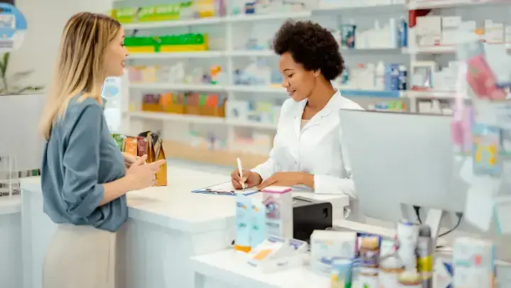 Zwei weibliche Personen in einer Apotheke: Eine steht hinter dem Bedientresen in einem weißen Kittel und notiert gerade etwas, die andere steht mit hellem Rock und dunkelgrüner Bluse bekleidet vor dem Tresen.