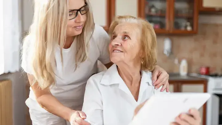 Eine Frau legt von schräg hinten ihre Hände vertraulich auf die Schultern einer älteren Frau. Die ältere Frau sitzt, sie zeigt der jüngeren Frau etwas auf einem Tablet.