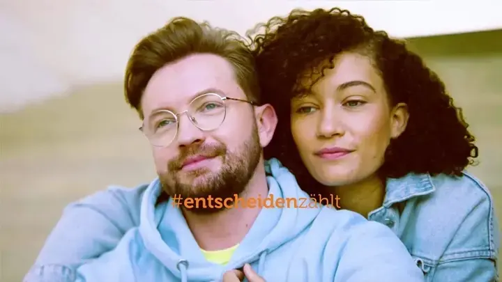 Eine Frau mit dunklen Locken umarmt einen Mann mit blonden Haaren und Bart von hinten. Beide schauen an der Kamera vorbei, Richtung Himmel. Slogan: #entscheidenzählt