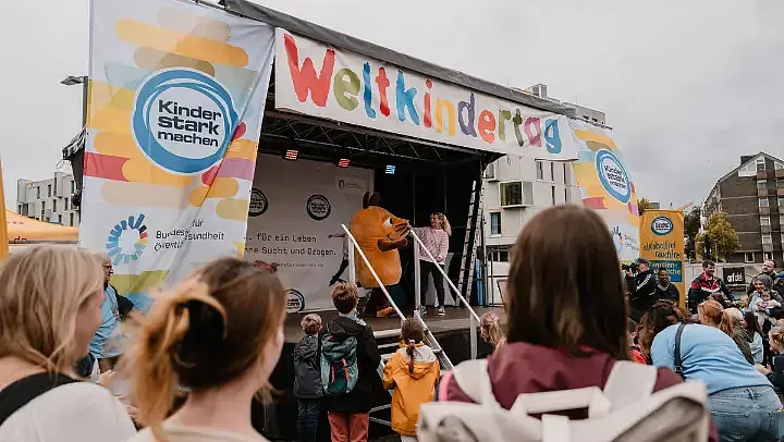 Bühne der Initiative “Kinder stark machen“ auf dem Weltkindertagsfest in Köln 2025. Auf der Bühne ist die Moderatorin Singa mit der Maus. Im Vordergrund sind Besucherinnen und Besucher von hinten zu sehen.