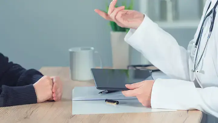 Beratungssituation: Zwei Menschen sitzen sich gegenüber an einem Tisch. Von der linken Person sieht man nur die Hände und von der rechten Person Hände, Unterarme und einen Teil des Oberkörpers. Daran kann man erkennen, dass es sich medizinisches Personal handelt. Die Person hat ein Stethoskop um den Hals hängen und hält ein Tablet in der linken Hand.