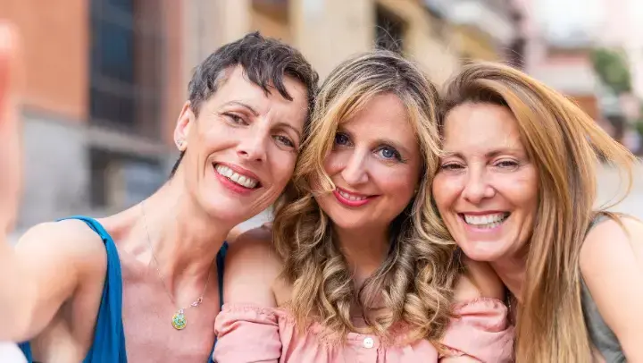 Drei Frauen mittleren Alters machen ein Selfie von sich. Im Hintergrund ist sind Häuserfassaden zu sehen.