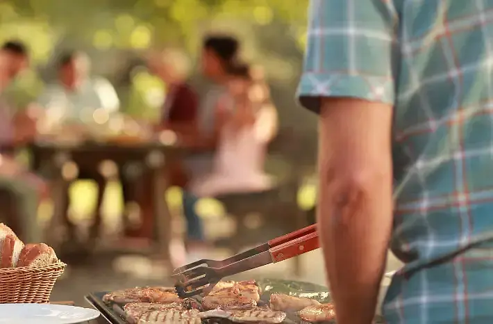 Eine Person grillt Fleisch auf einer Grillplatte. Im unscharfen Hintergrund sitzen mehrere Menschen gemeinsam an einem Tisch und essen. Die Szene vermittelt eine gesellige Grillfeier im Freien.