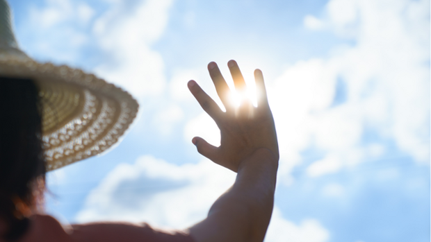 Person mit Sonnenhut von hinten im Anschnitt, die ihre Hand schützend in Richtung Sonne streckt