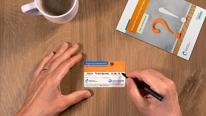 Draufsicht: Mittig im Bild ein Organspendeausweis, der von einer Hand gehalten und von der anderen Hand ausgefüllt wird. Oben links ist eine Kaffeetasse und oben rechts eine Broschüre zum Thema Organspende zu sehen.