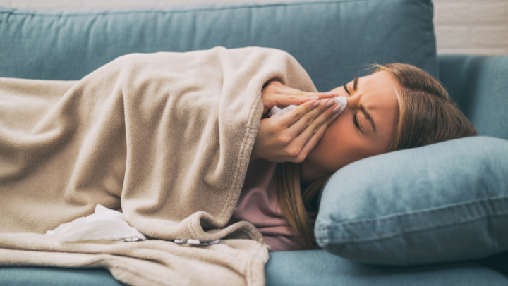 Eine Frau putzt sich die Nase und liegt auf dem Sofa unter einer Decke
