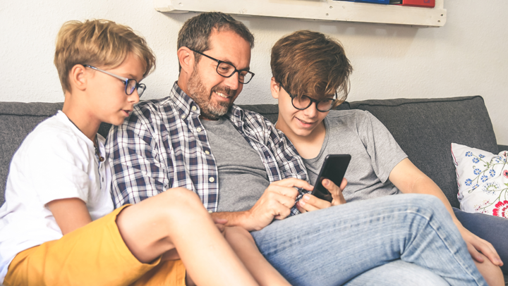 Ein Vater sitzt mit seinen zwei Teenager-Söhnen auf dem Sofa und sie schauen in ein Smartphone