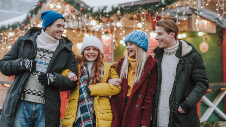 Vier junge Erwachsene stehen winterlich gekleidet eng beieinander. Im Hintergrund steht ein weihnachtlich geschmücktes Holzhaus. Alle lachen und wirken fröhlich.