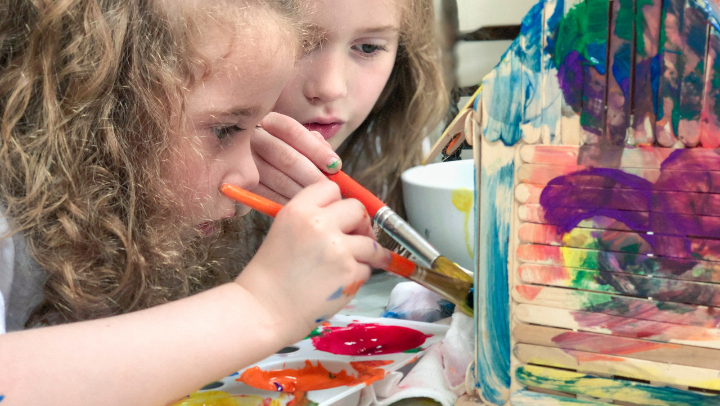 Zwei langhaarige Kinder malen konzentriert ein Holzhaus mit Pinseln an
