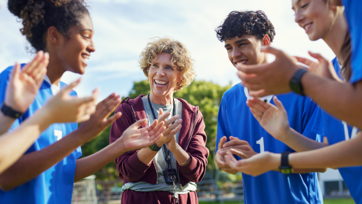 Trainerin (in der Mitte des Bildes) motiviert um sie herum im Kreis stehende Jugendliche. Alle klatschen in die Hände.