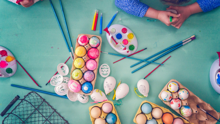 Bunt bemalte Eier in Eierkartons und Pinsel sowie Kinderhände aus der Vogelperspektive