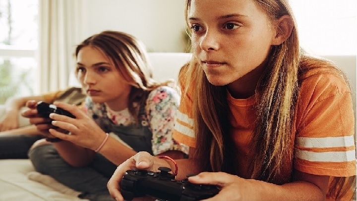 Zwei jugendliche Mädchen liegen auf dem Bauch und halten Controller in der Hand. Sie schauen seitlich an der Kamera vorbei, vermutlich auf einen Bildschirm.