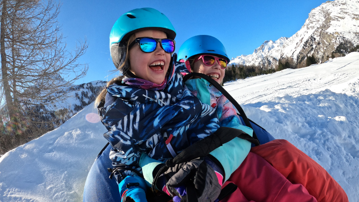 Zwei Kinder sitzen lachend auf einem Schlitten in einer verschneiten Landschaft. Beide tragen Helme und Sonnenbrillen.