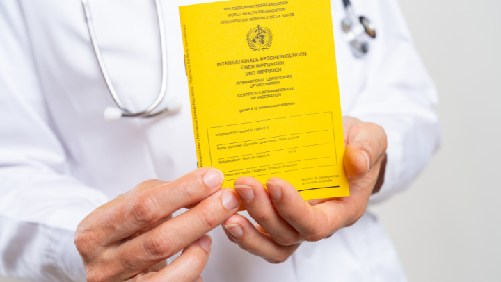 Im Vordergrund: Hände, die einen Impfpass ins Bild halten; im Hintergrund ein Mensch in weißem Kittel, mit Stetoskop um den Hals