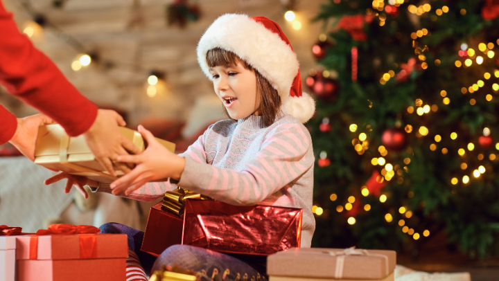 Mädchen mit Weihnachtsmütze sitzt vor dem Weihnachtbaum und erhält Geschenke