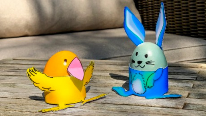 Zwei bunte Ostereier, ein gelbes und ein grünes, in gebastelten Papiereierbechern. Der gelbe sieht aus wie ein Küken, der blaue wie ein Hase. 