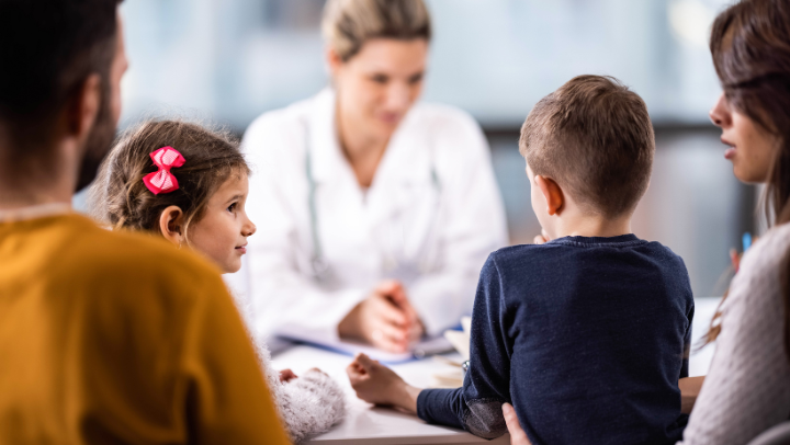 Familie (Vater, Tochter, Sohn, Mutter) im Gespräch mit einer Ärztin. Die Familie (scharf im Vordergrund) ist von hinten zu sehen, die Ärztin von vorne, allerdings unscharf im Hintergrund.