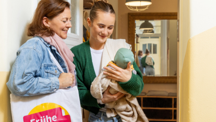 Zwei Frauen stehen nebeneinander in einem Flur. Eine hält ein Baby im Arm. Beide Frauen blicken lächelnd auf das Baby.