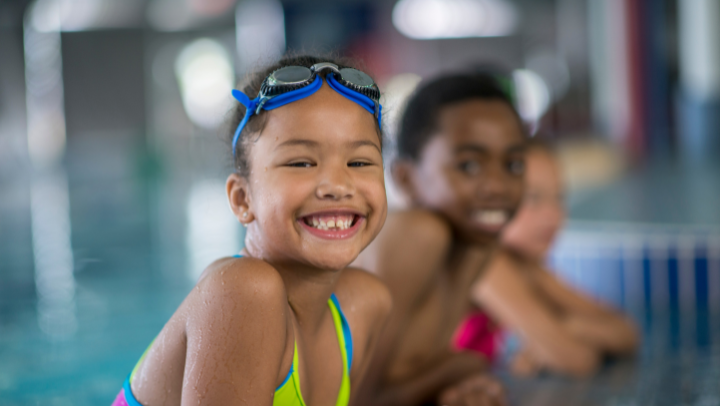 Ein Mädchen im Badeanzug und mit auf die Stirn gezogener Schwimmbrille stützt sich auf der Umrandung eines Pools ab und lächelt in die Kamera. Im Hintergrund sind verschwommen zwei weitere Kinder zu sehen.