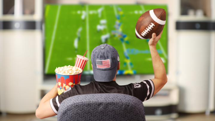 Junger Mann von hinten zu sehen: Sitzt mit Popcorn, Getränk und Football vor einem großen Fernseher, auf dem unscharf ein Footballspiel zu sehen ist.