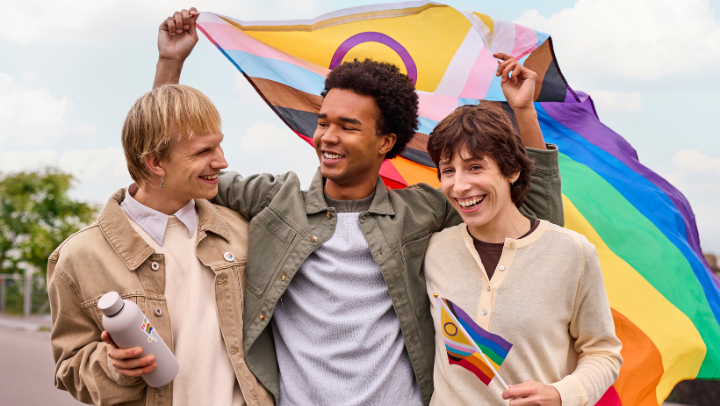 Drei junge Personen Arm in Arm, lachen fröhlich. Die mittlere hält in beiden Händen eine Pride-Flagge hinter sich hoch.