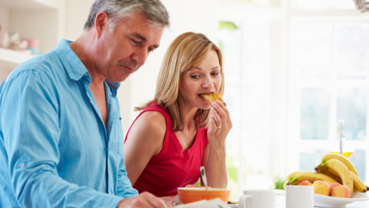Mann und Frau mittleren Alters sitzen nebeneinander, Mann liest Zeitung, Frau isst ein Brot. Beide schauen auf die Zeitung.