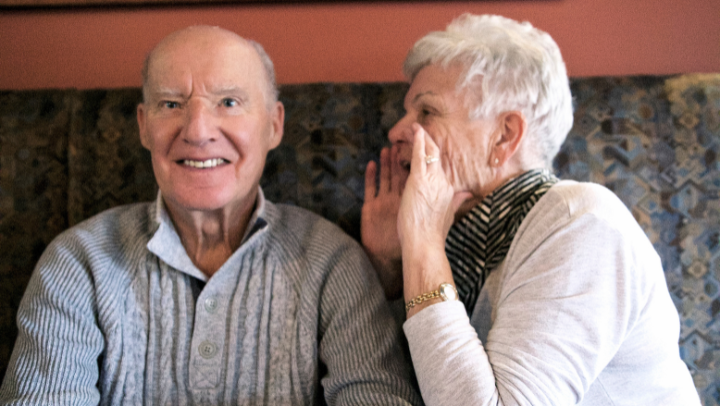 Ein älterer Mann und eine ältere Frau sitzen auf einem Sofa. Der Mann blickt Richtung Kamera, die Frau hält ihre Hände zu einem Trichter geformt an ihren Mund und schreit in Richtung des Mannes.