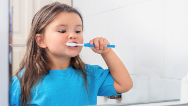 Mädchen in blauem T-Shirt steht vor dem Spiegel und putzt sich die Zähne.