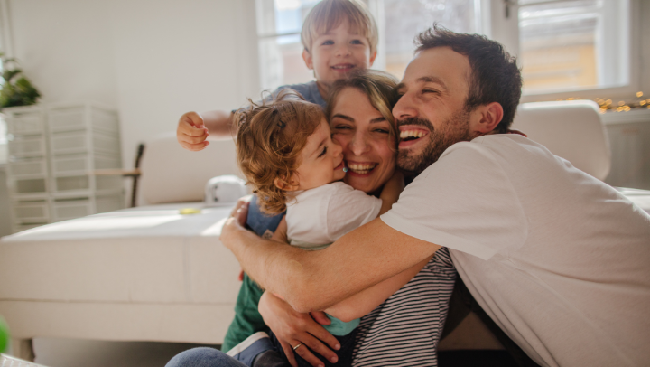 Familie – Mama, Papa, Sohn und Tochter – umarmen sich lachend auf dem Boden vor einem weißen Sofa. 