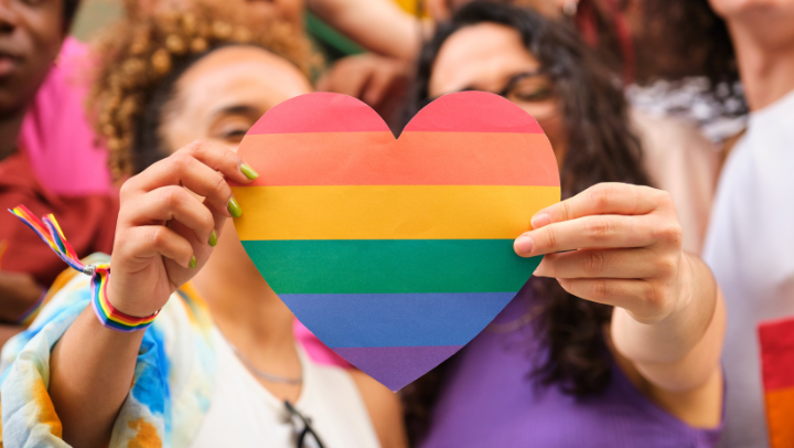 Zu sehen ist ein Herz in Regenbogenfarben. Es signalisiert Vielfalt und wird von zwei Frauen in den Händen gehalten. 