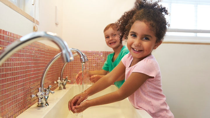 Zwei Kinder stehen an einem großen Waschbecken mit drei Wasserhähnen und halten ihre Hände unter fließendes Wasser. Beide lächeln in Richtung der Kamera.
