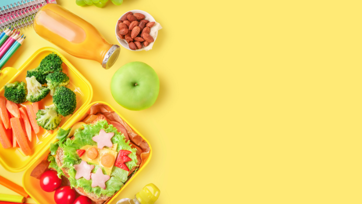 Auf einem gelben Untergrund liegen Stifte, Ringblöcke, ein Apfel, ein Schälchen mit Mandeln und eine geöffnete Brotdose, die Gemüsesorten und ein belegtes Brot enthält.