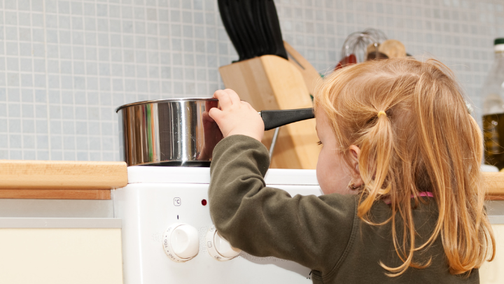Kind mit langen Haaren steht mit dem Rücken zur Kamera an einem Herd und berührt einen Topf, der darauf steht.