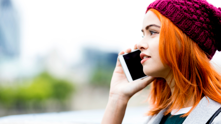 Junge Frau mit orangefarbenen Haaren und dunkelroter Strickmütze hält ein Handy ans Gesicht.