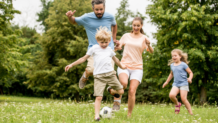 Familie spielt gemeinsam Fußball auf einer Wiese im Grünen; die Kinder laufen lachend vor den Eltern her.