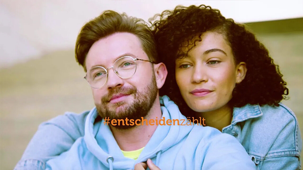 Eine Frau mit dunklen Locken umarmt einen Mann mit blonden Haaren und Bart von hinten. Beide schauen an der Kamera vorbei, Richtung Himmel. Slogan: #entscheidenzählt