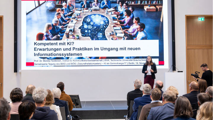 Auf einer Bühne steht eine Frau und spricht vor einem sitzenden Publikum. Hinter ihr ist eine große Präsentationsfolie mit dem Titel „Kompetent mit KI? Erwartungen und Praktiken im Umgang mit neuen Informationssystemen“. Auf der Folie ist ein großes Bild von Menschen zu sehen, die an einem langen Tisch sitzen, darüber grafische Darstellungen eines Gehirns und technischer Begriffe. Neben der Bühne befindet sich ein Mann mit einer Kamera.